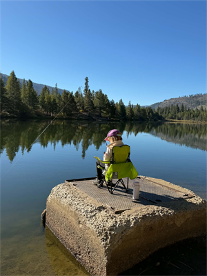 A young girl sitting and fishing in a green folding chair on top of a concrete block in a lake.