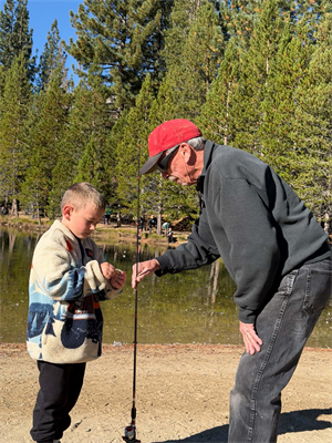 An old man holding a fishing pole for a young boy to bait in front of a lake.